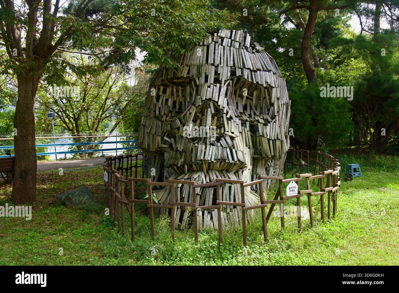 Una grande scultura in legno con testa intemprata sorge dietro una bassa recinzione nel boschetto ombreggiato del parco del Museo d'animazione di Chuncheon. Foto Stock