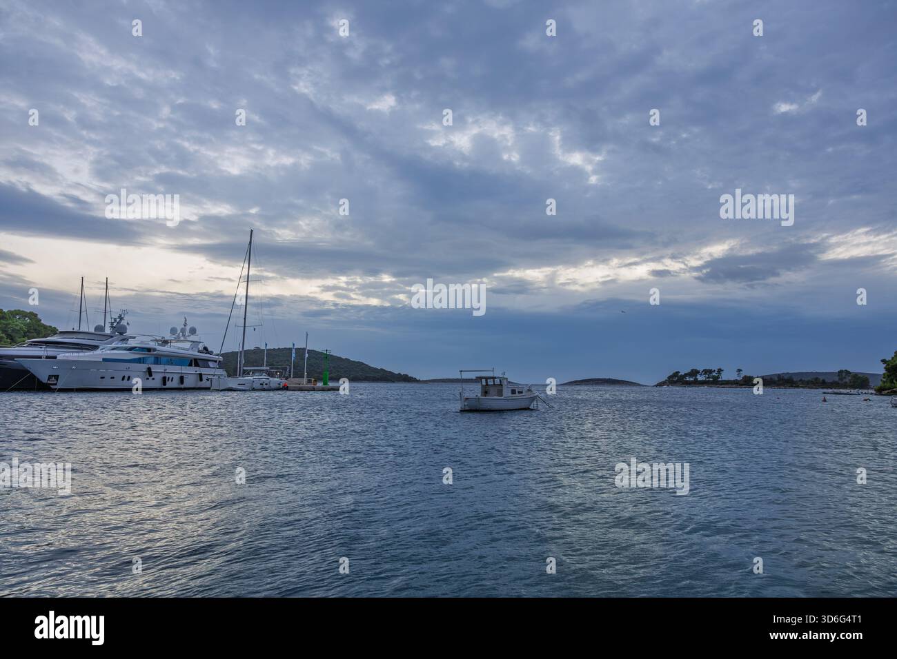 Un tranquillo porto con grandi yacht di lusso attraccati a sinistra e una piccola barca solitaria che attraversa acque calme sotto un cielo blu. Ideale per la nautica Foto Stock