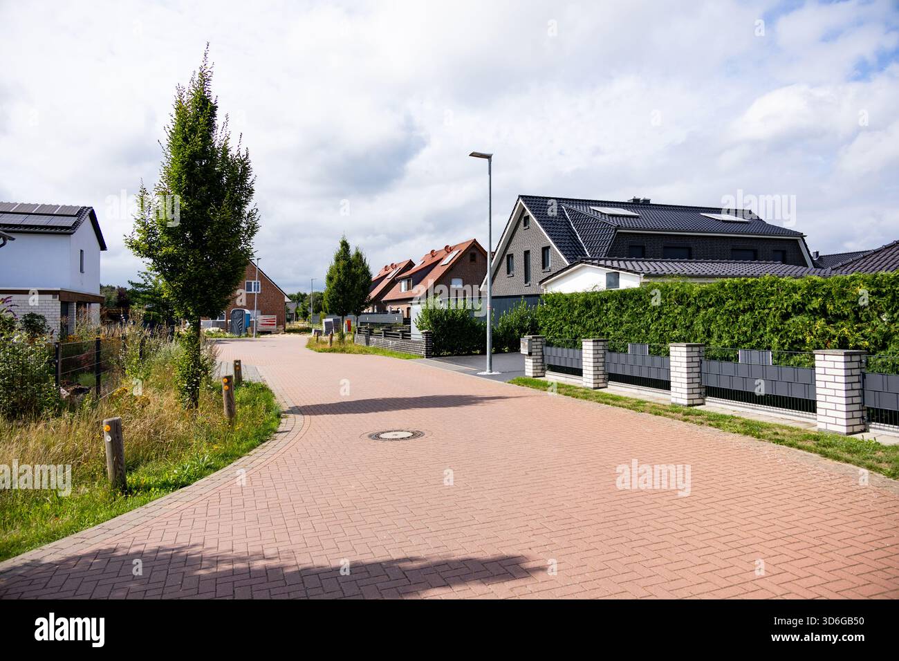 Tranquilla strada residenziale in un moderno quartiere periferico con case di nuova costruzione, verde e un passaggio pedonale pavimentato sotto un cielo parzialmente nuvoloso Foto Stock