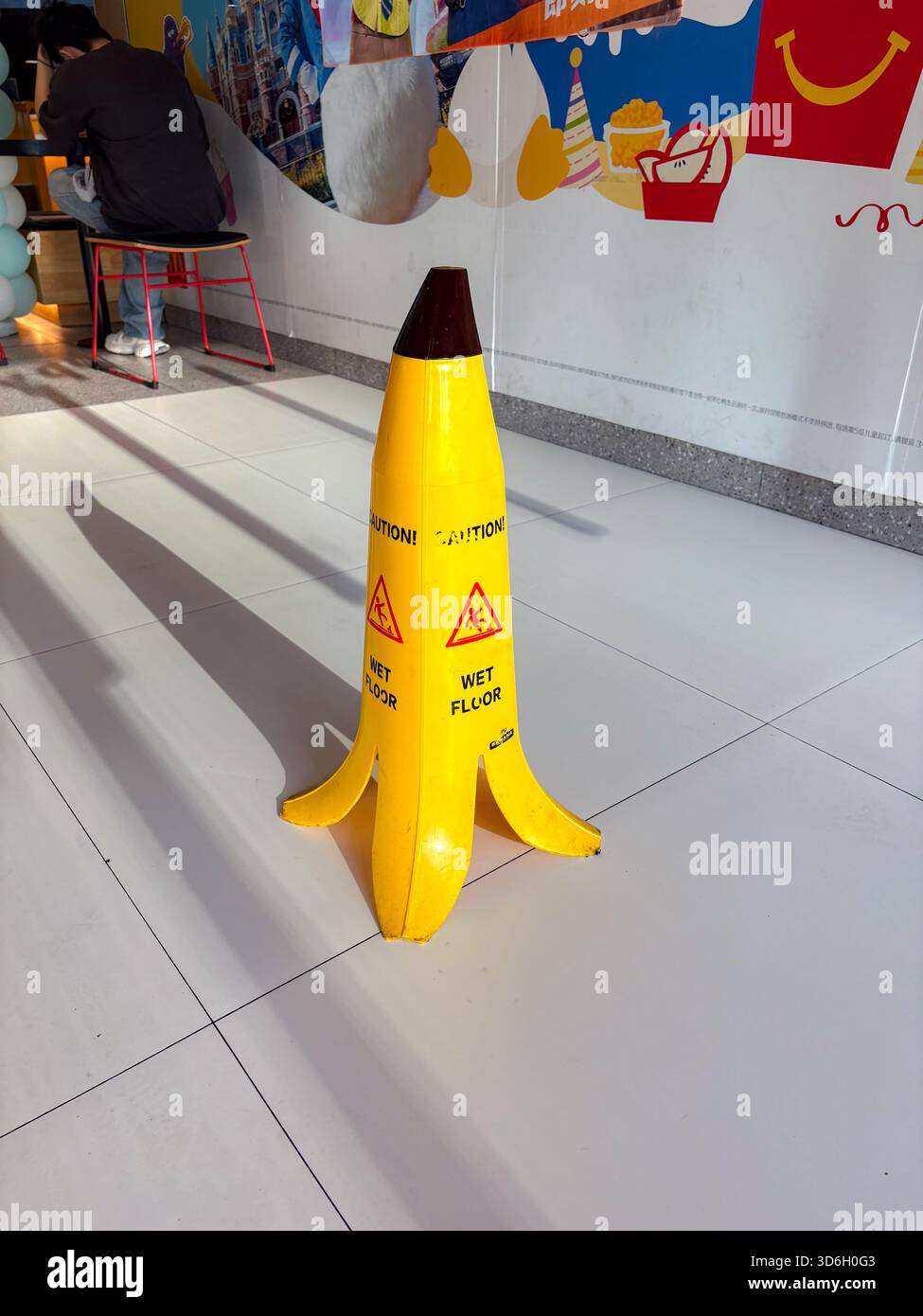 Cartello a strappo per pavimenti umidi con buccia di banana. Un esclusivo cono di avvertenza giallo si trova su un pavimento piastrellato pulito, con simboli di attenzione per le aree umide. Divertente ma funzionale Foto Stock