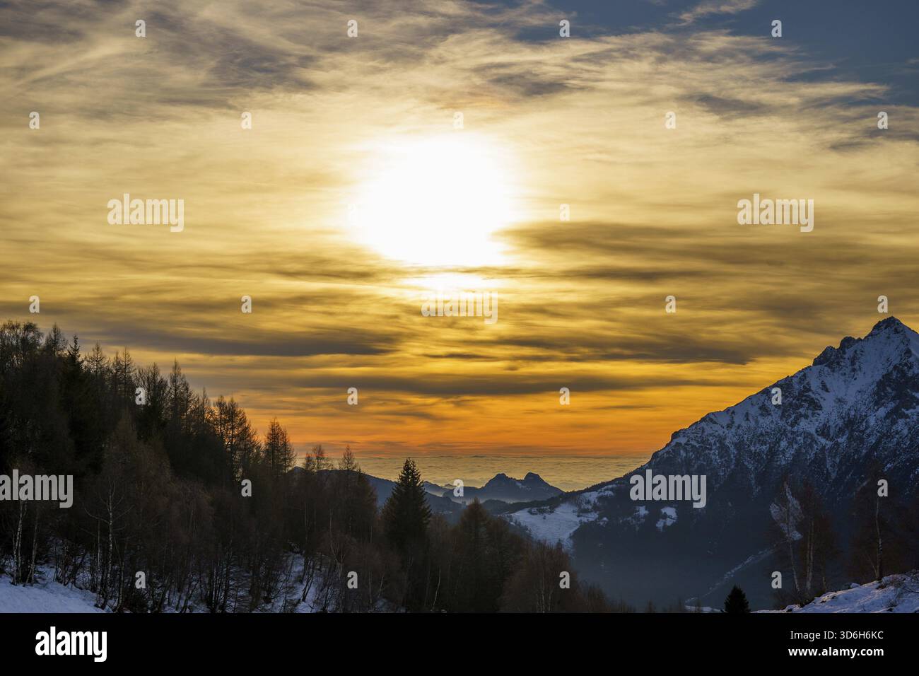 Il sole tramonta sulla cima innevata della montagna, che emette un caldo bagliore dorato sul cielo e crea un contrasto drammatico con l'aspro paesaggio alpino piani Foto Stock