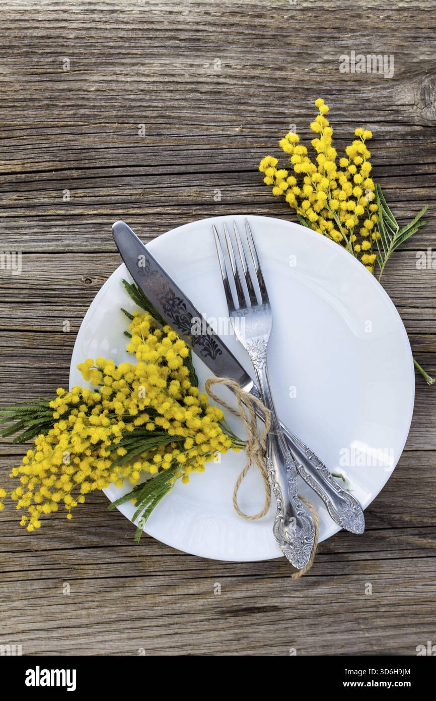 Cibo, coltello a forchetta con piatto e fiori di mimosa su un tavolo di legno per la cena delle vacanze di Pasqua di primavera. Vista dall'alto Foto Stock