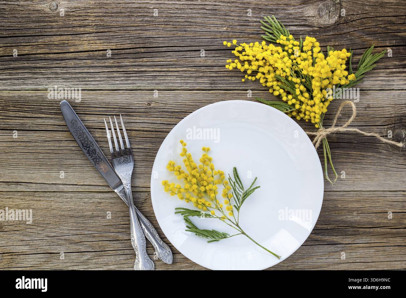 Cibo, coltello a forchetta con piatto e fiori di mimosa su un tavolo di legno per la cena delle vacanze di Pasqua di primavera. Vista dall'alto Foto Stock