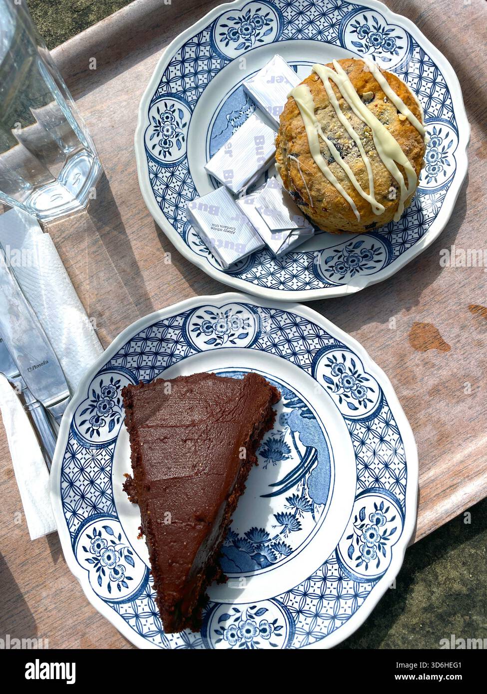 Fetta di ricca torta al cioccolato e ampio scone con spruzzino bianco servito su piatti tradizionali a motivi geometrici con porzioni di burro su un vassoio di legno uk Foto Stock