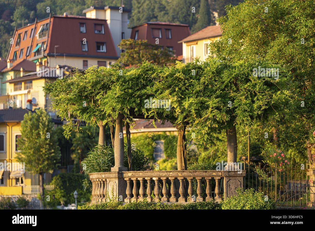 Lussureggiante pergola ricoperta di vegetazione sulla balaustra decorativa in pietra, adagiata sullo sfondo di case colorate del villaggio. Una luce calda migliora il summ vibrante Foto Stock