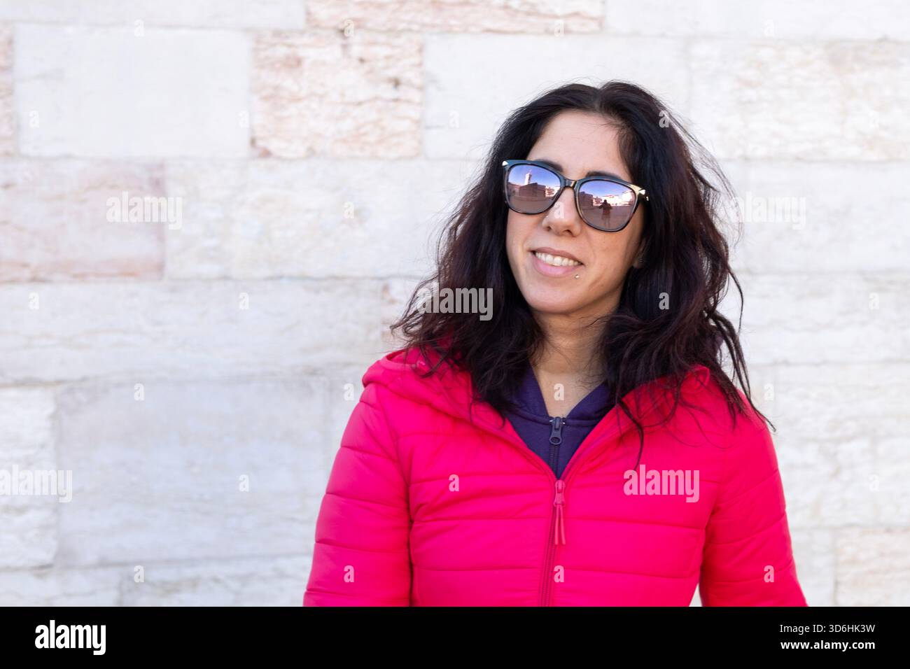 Donna dai capelli scuri che posa di fronte a un antico muro. La donna ha trentotto anni e indossa un piumino fucsia Foto Stock