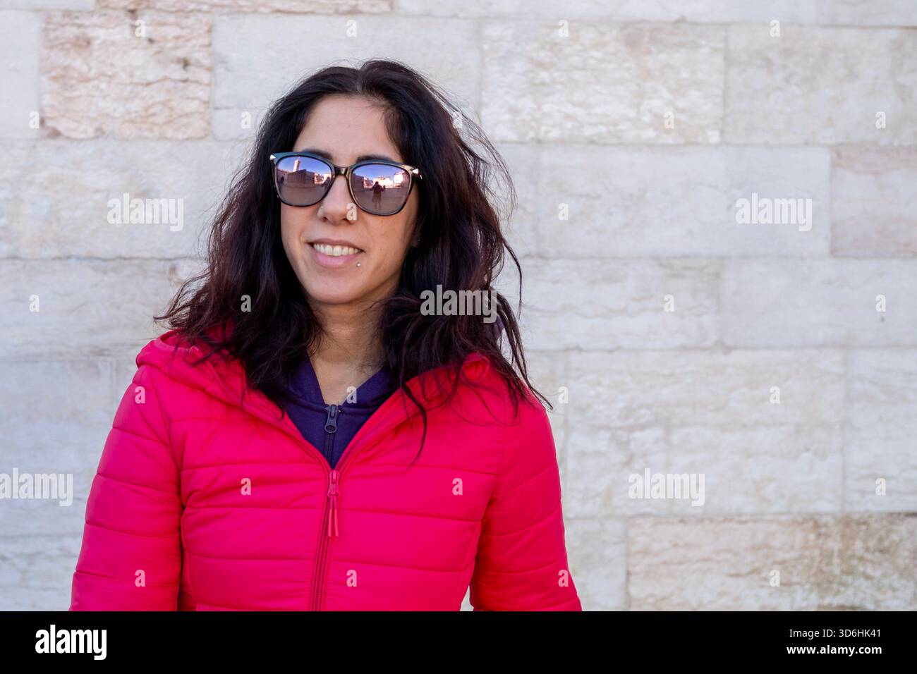 Donna dai capelli scuri che posa di fronte a un antico muro. La donna ha trentotto anni e indossa un piumino fucsia Foto Stock