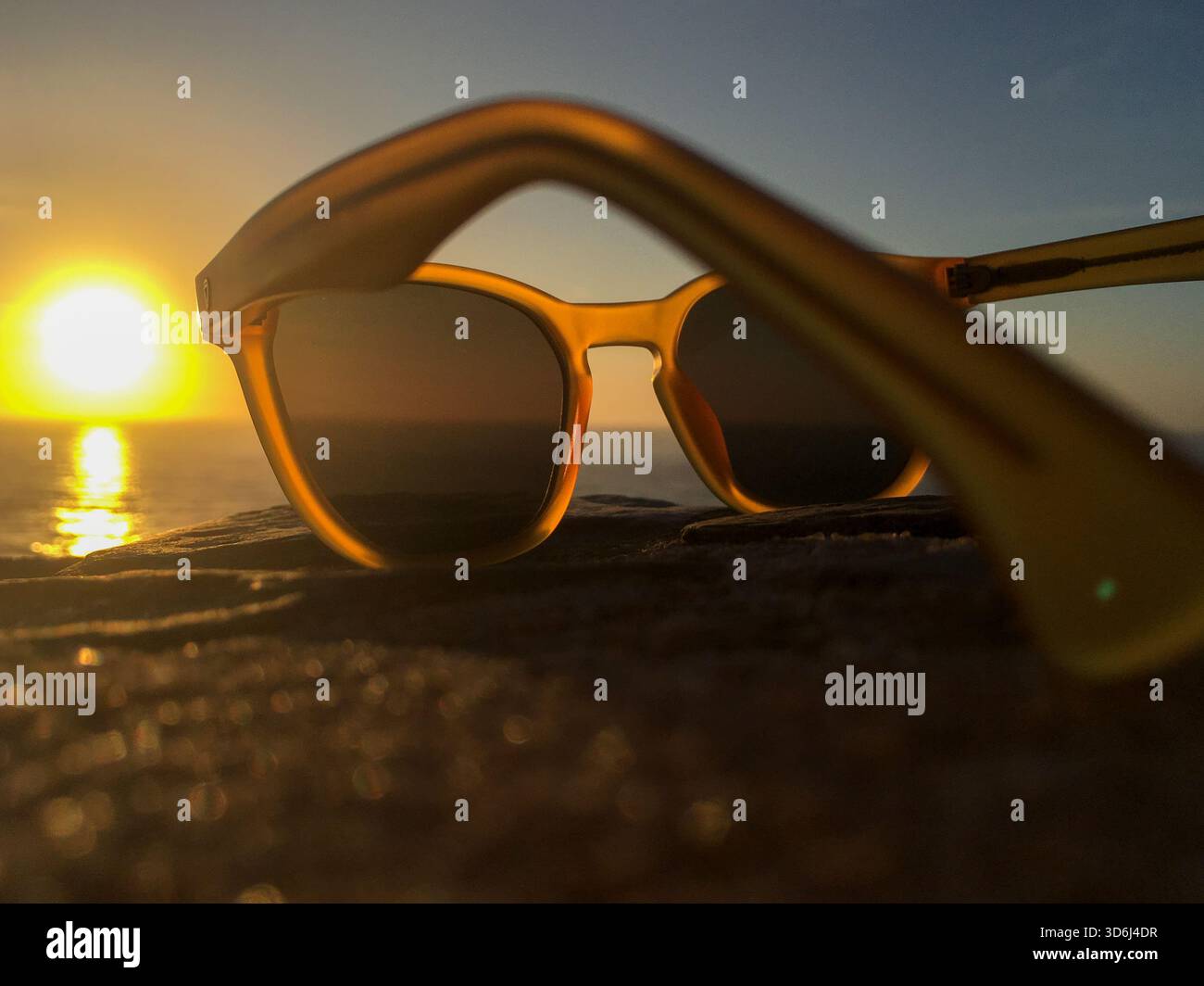 Primo piano di eleganti occhiali da sole posti all'aperto con splendida vista del tramonto sull'oceano. La luce dorata e soffusa crea un'atmosfera rilassata per l'estate e i viaggi. Foto Stock