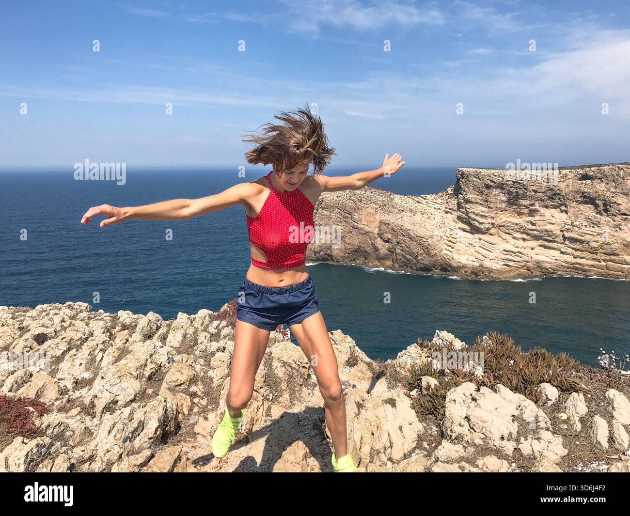 Donna sportiva che salta giocosamente sulle rocce costiere in una giornata di sole vicino a Sagres, Portogallo. Oceano Atlantico alle sue spalle, mentre ama viaggiare e fare visite turistiche. Foto Stock