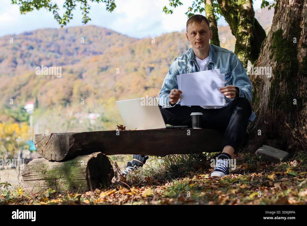 Uomo all'aperto su un tronco di legno simile a una panchina, che espone fogli vuoti, computer portatile e caffè presenti in un vivace scenario autunnale Foto Stock