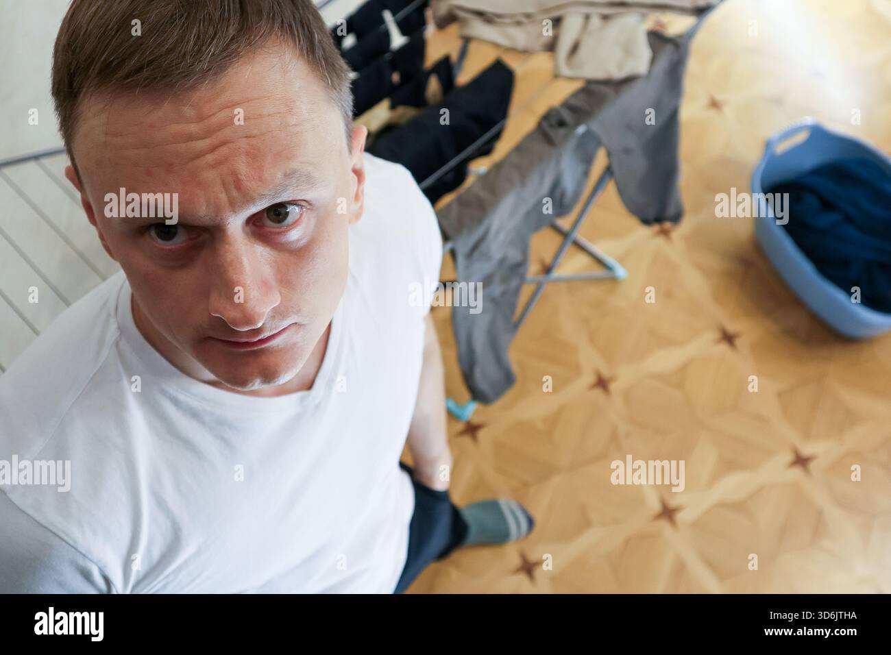 Primo piano di un giovane frustrato con una t-shirt bianca che guarda verso l'alto in un'area lavanderia con vestiti su stendino e cesto nelle vicinanze Foto Stock