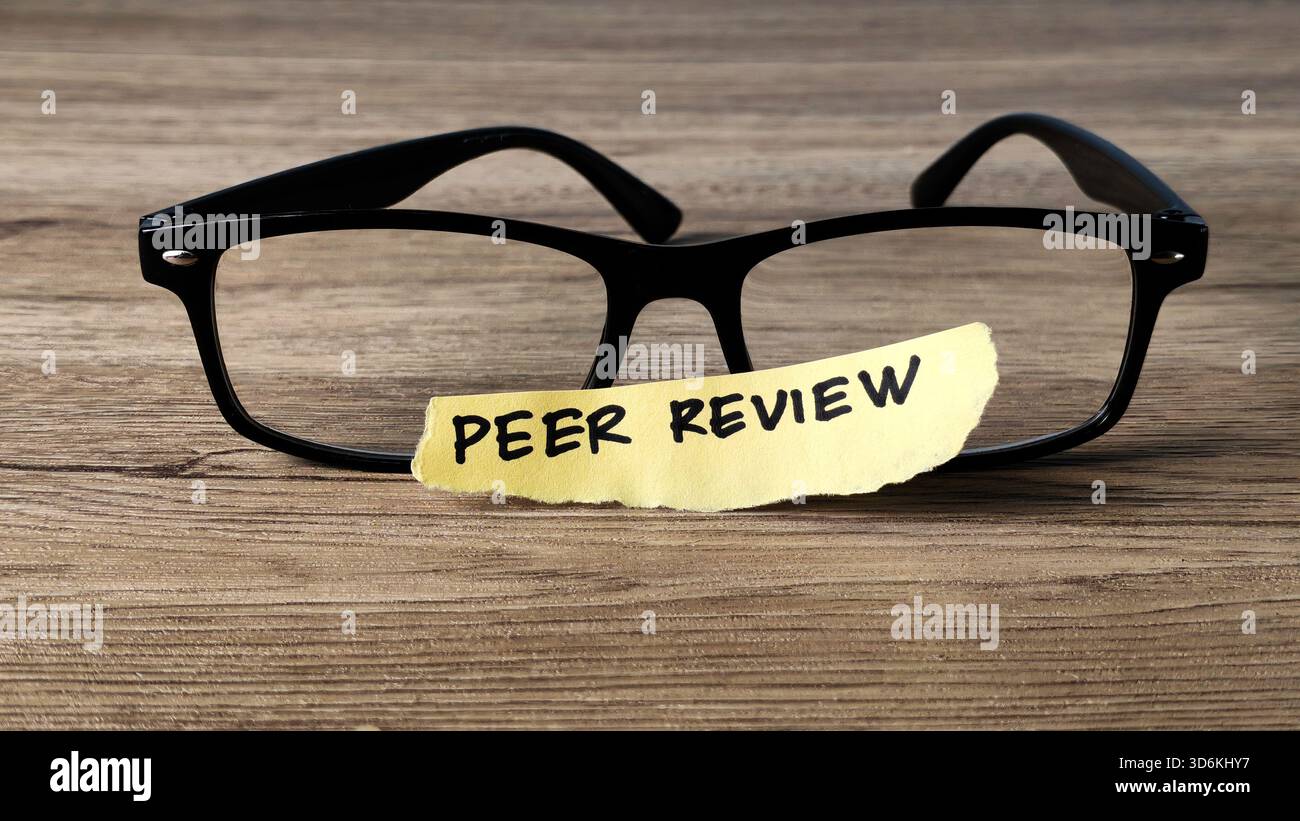 Concetto di revisione inter pares con una nota scritta a mano posizionata su occhiali da lettura su una scrivania di legno, che simboleggia la ricerca accademica e la garanzia della qualità. Foto Stock