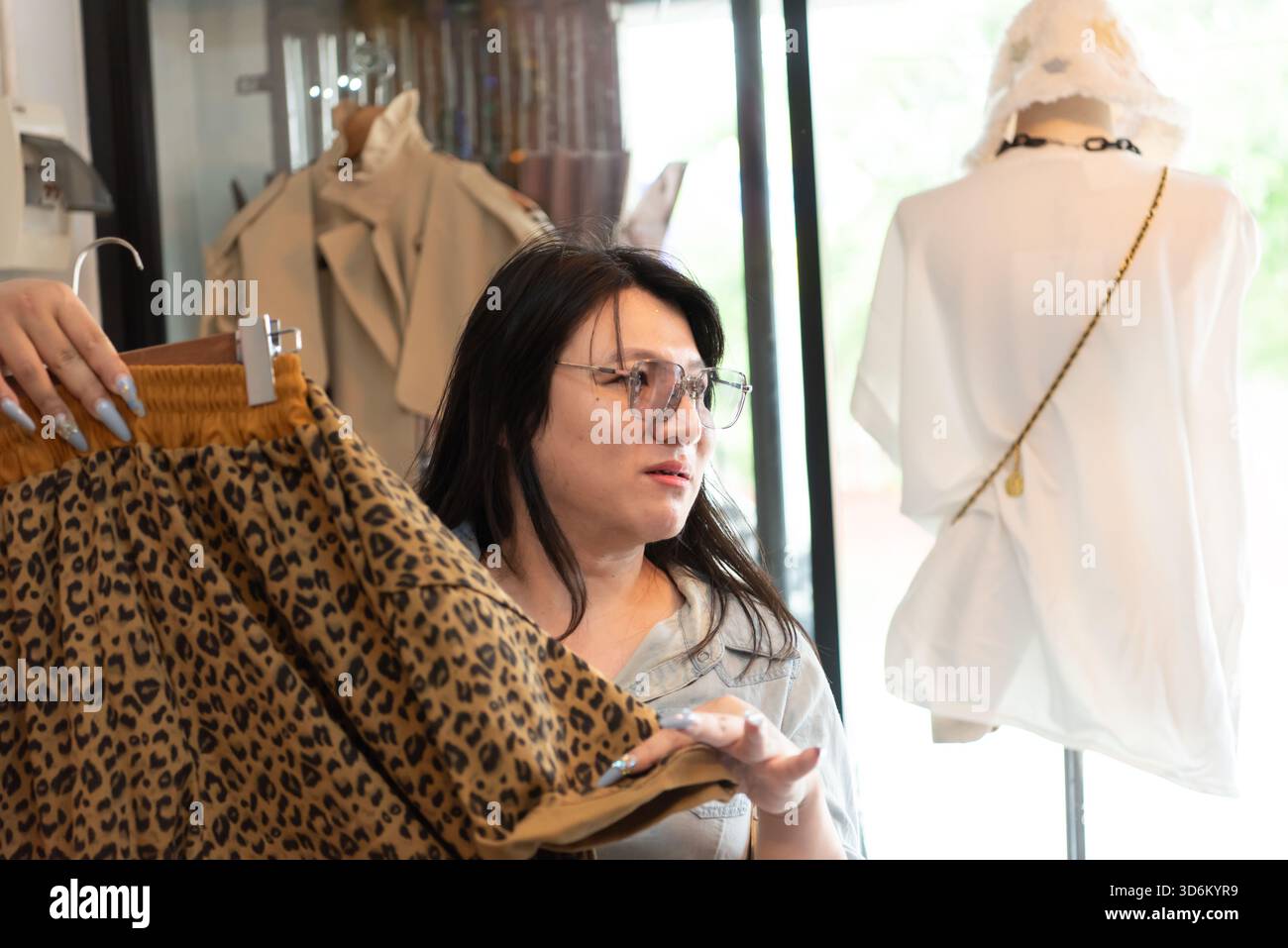 Due amici LGBTQ si divertono a fare shopping in un negozio alla moda. Uno regge la gonna con stampa leopardata, discutendo delle opzioni. Il negozio presenta Moder Foto Stock