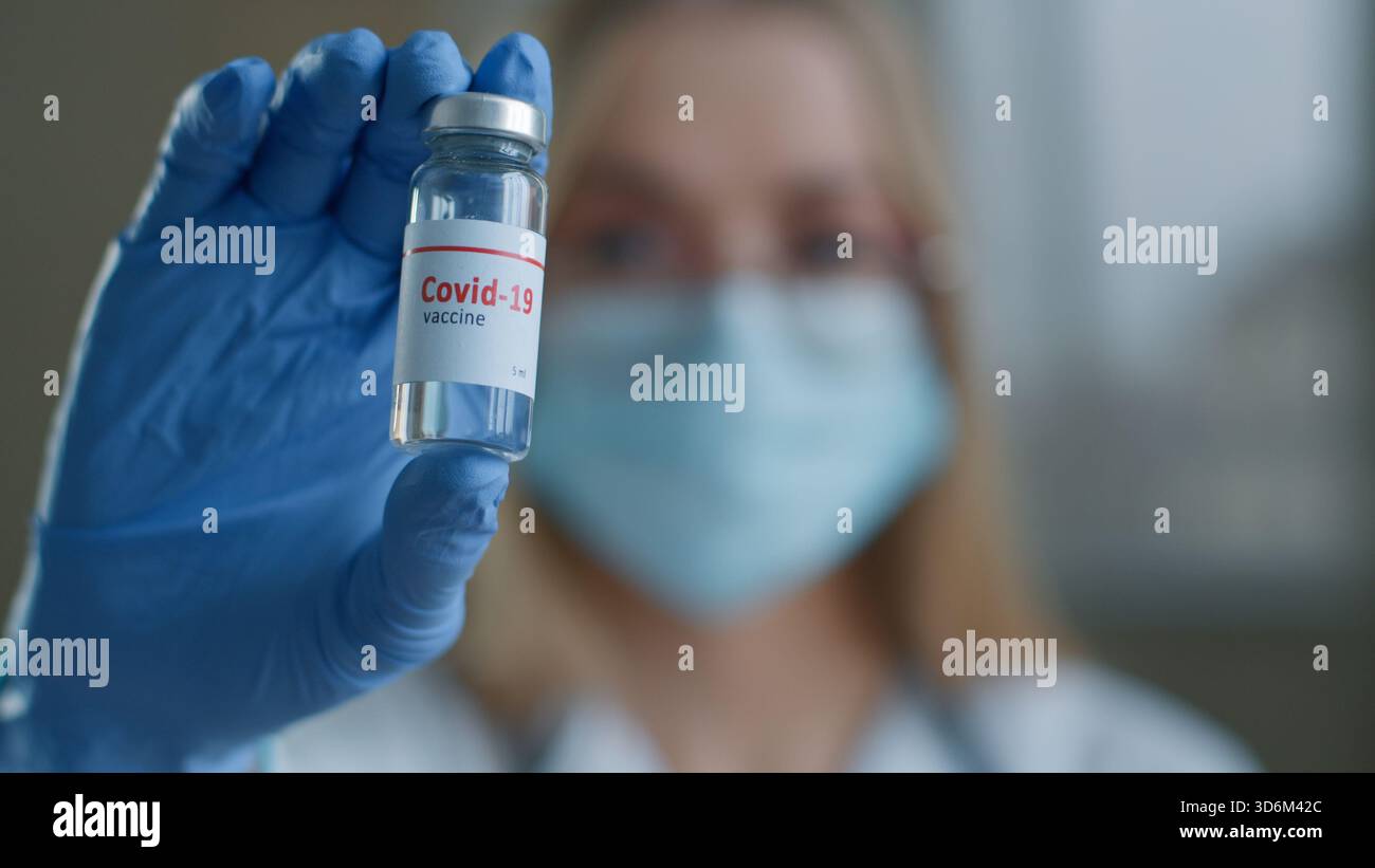 Primo piano donna medico che mostra vaccino iniezione bottiglia in laboratorio scienziata infermiera ragazza in maschera medica guardando la fotocamera che tiene covid19 Foto Stock