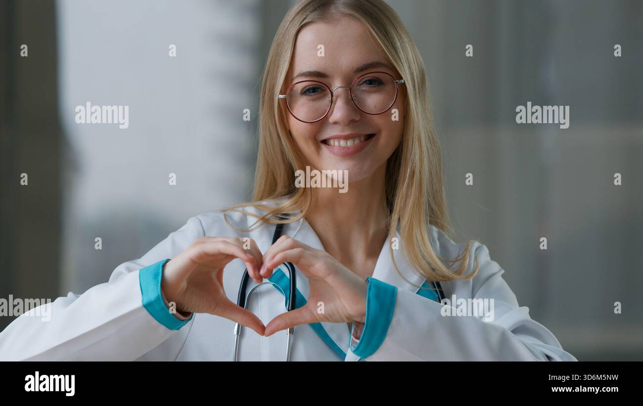 Donna sorridente caucasica medico che mostra forma cuore cardiologa infermiera ragazza femminile mostrare gesto cardio guardando la macchina fotografica terapeuta Foto Stock