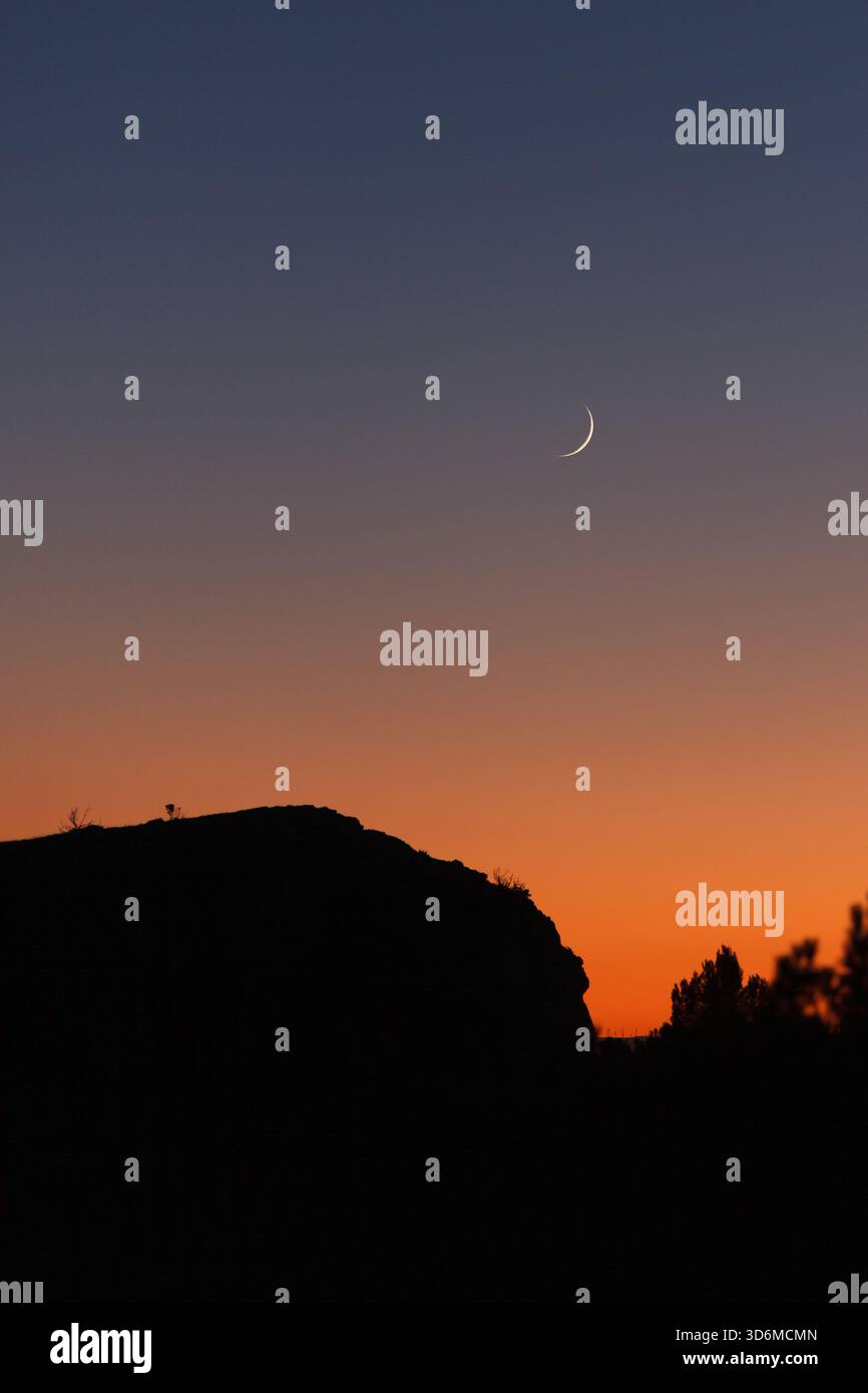 Cielo al tramonto minimalista con una sottile luna crescente sopra un caldo orizzonte arancione e un paesaggio dalle linee scure. Foto Stock