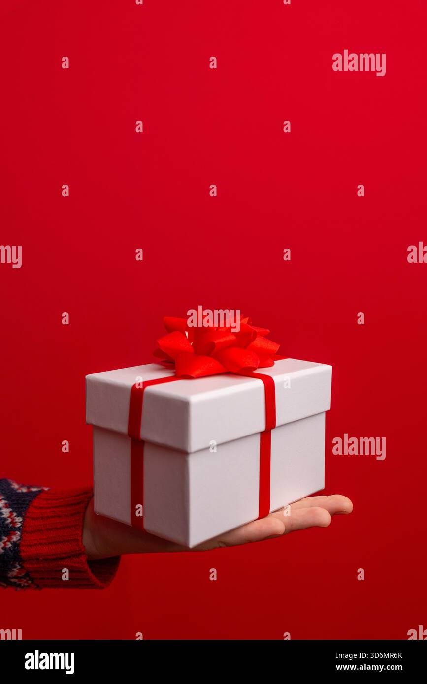 Confezione regalo natalizia con un fiocco rosso presentato sulla mano di un bambino, con un vivace sfondo rosso e accoglienti dettagli del maglione invernale Foto Stock