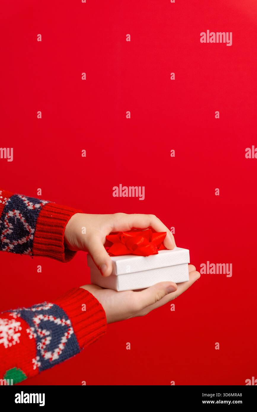Mani che tengono in mano una confezione regalo bianca chiusa con fiocco rosso su sfondo rosso, perfetta per layout di spazio, annunci natalizi e progetti promozionali Foto Stock