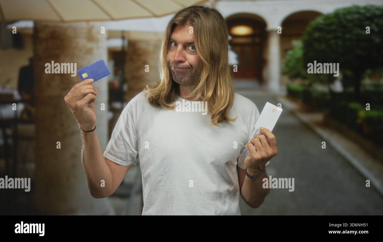 L'uomo biondo ha una carta di credito blu e una carta bianca in un edificio storico del chiostro universitario, alzando entrambe le mani con un'espressione sconcertata; decisione Foto Stock