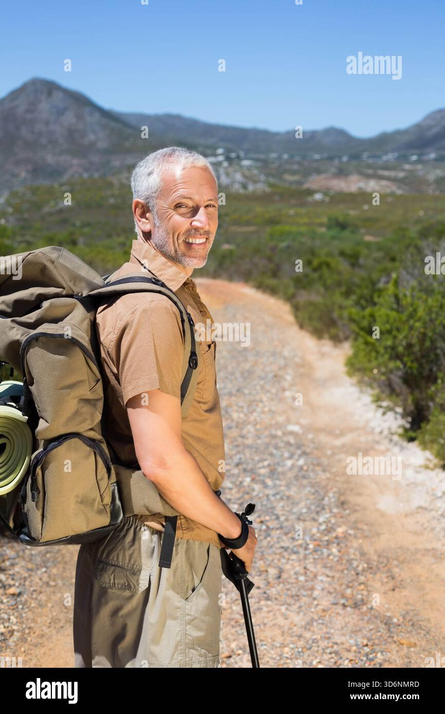 Uomo anziano in piedi sul sentiero di montagna guardando indietro, zaino di olive, pattino arrotolato legato, palo di tenuta. Escursionista, zaino, robusto, arido, paesaggio, viaggiare, o Foto Stock
