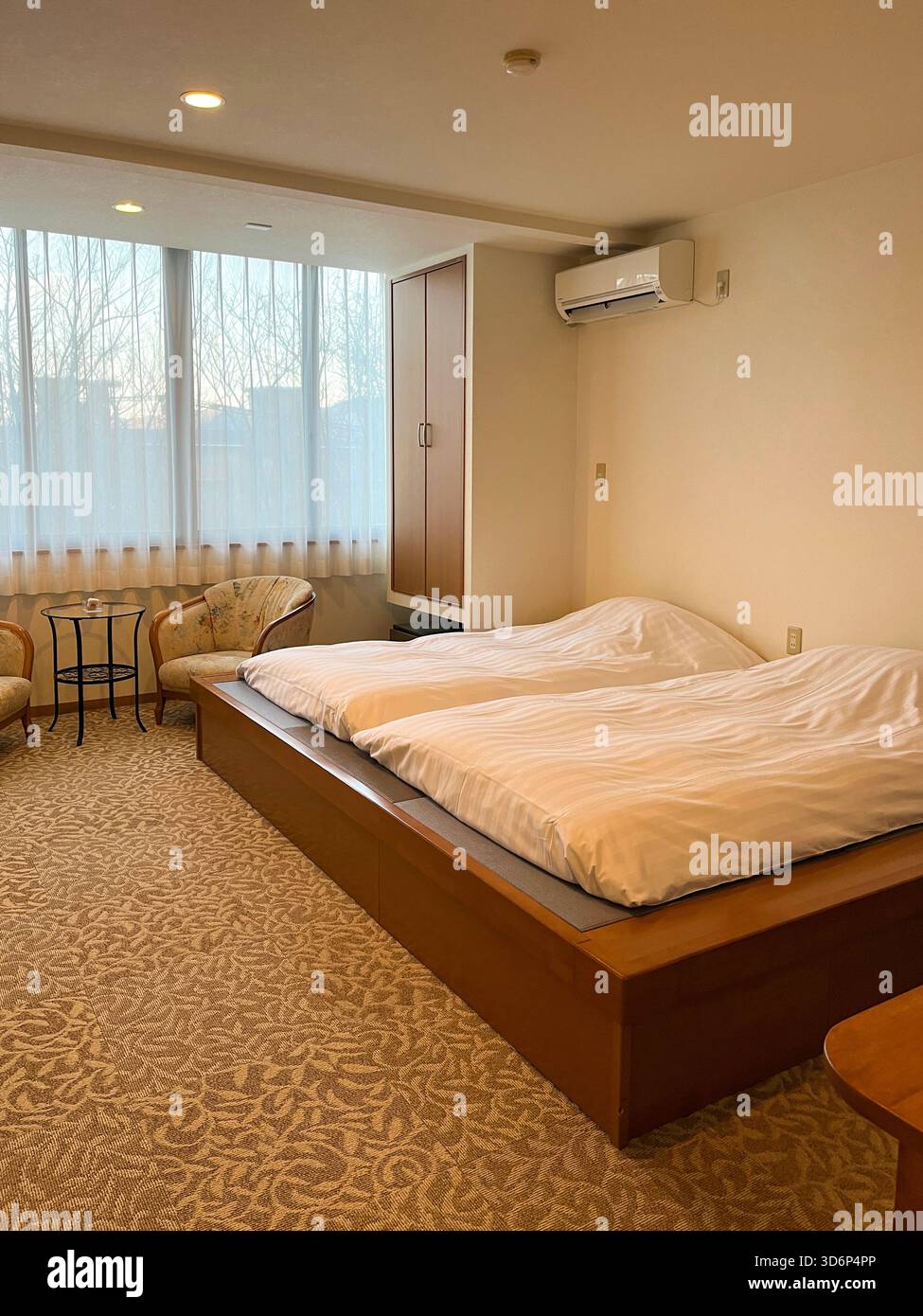 Camera intima in un tradizionale hotel ryokan giapponese, con bassa piattaforma in legno, letti gemelli, illuminazione soffusa, poltrone e grande finestra con curtai puro Foto Stock