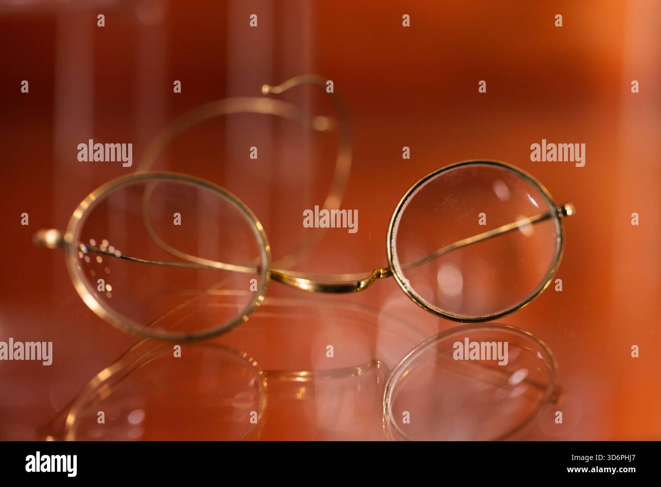 Gli eleganti occhiali rotondi vintage con sottili montature dorate poggiano su una superficie lucida e dai toni caldi. I riflessi morbidi creano una suita d'atmosfera contemplativa e senza tempo Foto Stock