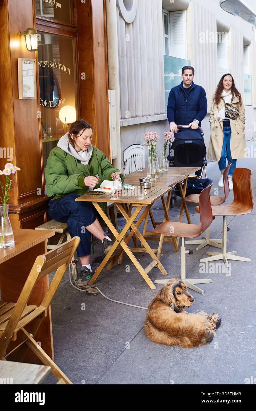 Parigi, Francia - marzo 2025: Una donna siede a un tavolino a mangiare, mentre una coppia con un passeggino si trova nelle vicinanze. Un cane si rilassa per terra all'esterno di Foto Stock