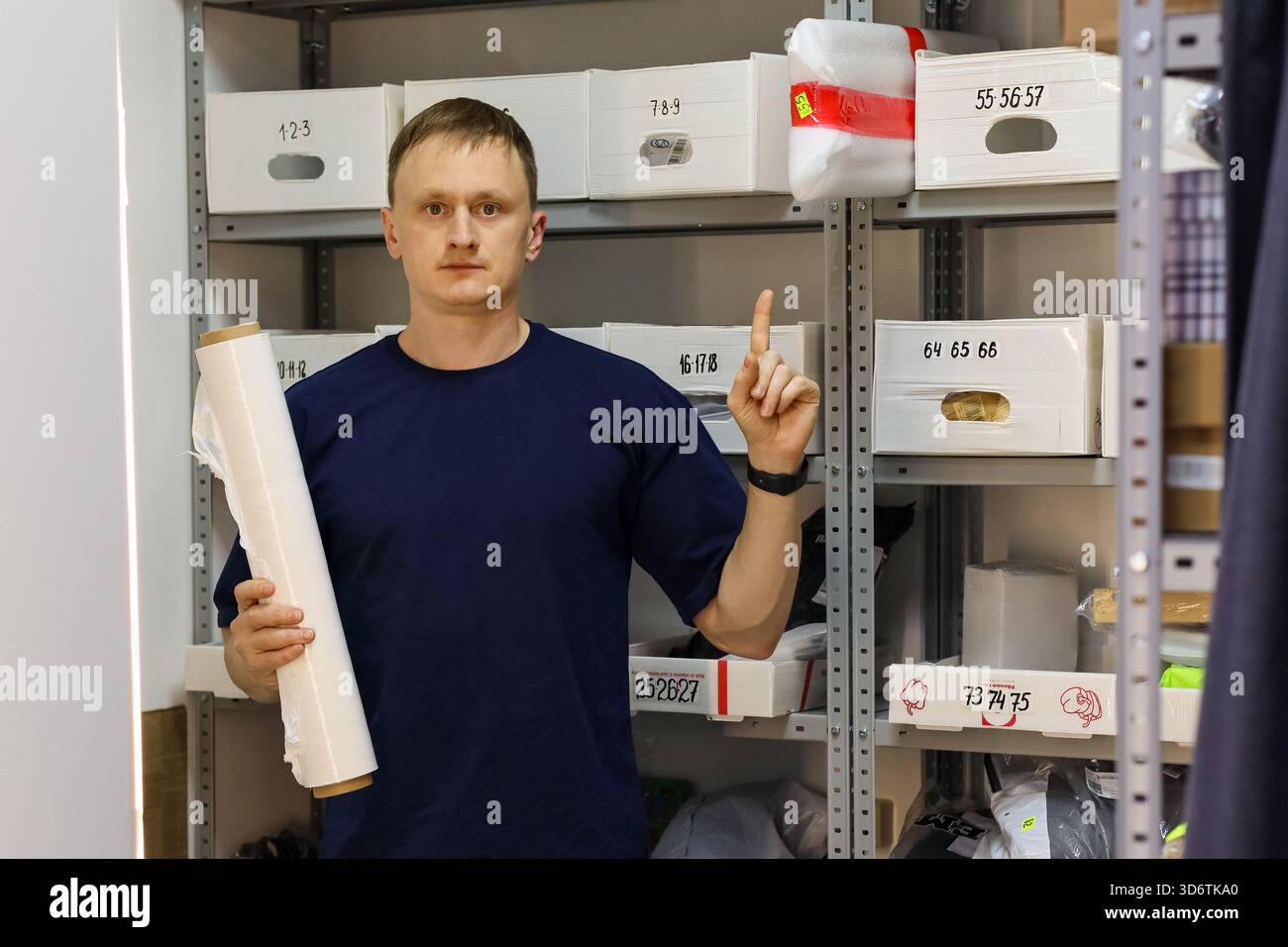 Il lavoratore con la camicia scura punta verso l'alto fino alla scatola etichettata sul ripiano metallico, tenendo in mano il rotolo bianco. Foto Stock