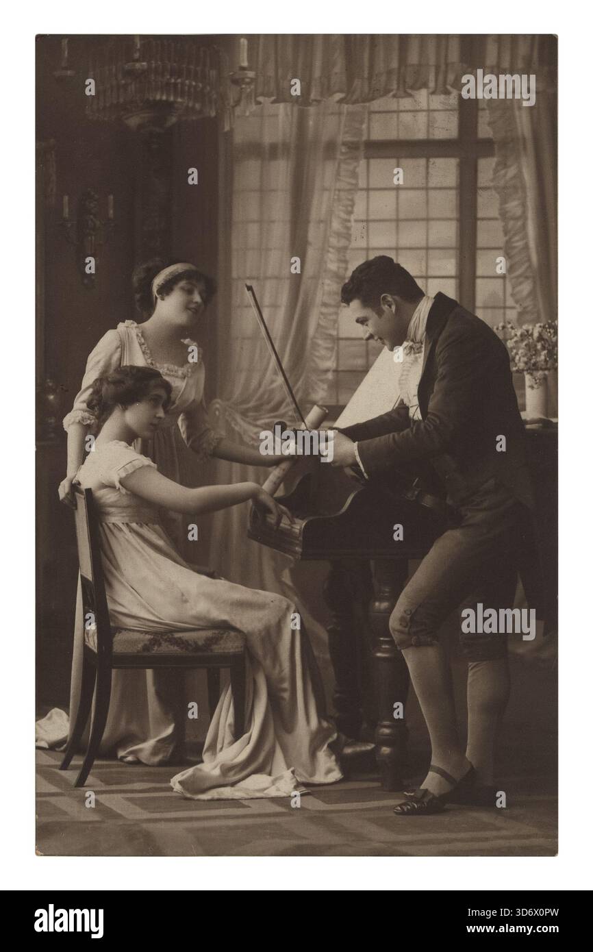 Una cartolina vintage dai toni seppia mostra tre persone in un salotto; un uomo suona il violino, una donna siede ad ascoltare un pianoforte e un'altra donna sta Foto Stock