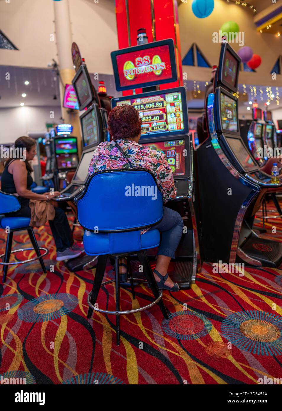 Donna che gioca a una luminosa slot machine all'interno di un casinò colorato, con tappeti a motivi geometrici, posti a sedere blu, schermi illuminati e una vivace atmosfera da gioco Foto Stock