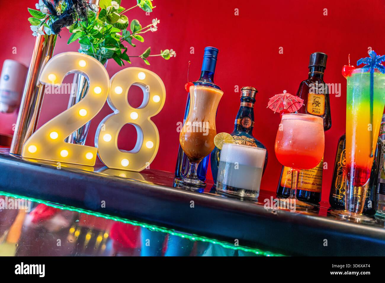 Cocktail colorati, bottiglie di liquori, caramelle ed elementi decorativi disposti su un tavolino da bar festivo con vibrante illuminazione di sfondo rosso. Foto Stock