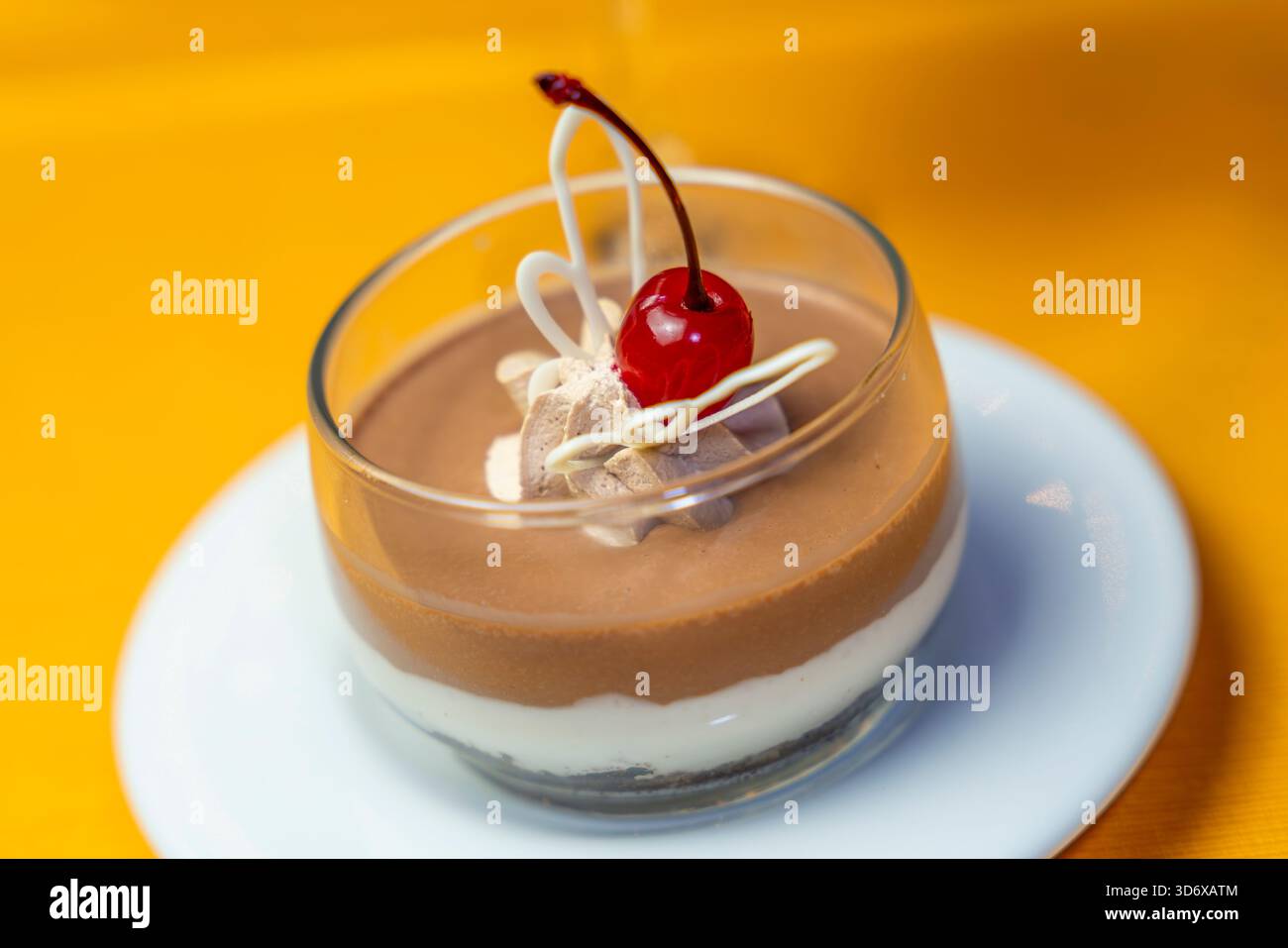 Dessert di panna cotta al cioccolato ricoperto di ciliegia, panna montata e decorazione di cioccolato bianco servito in una tazza di vetro su un piatto bianco su un coagulo giallo Foto Stock
