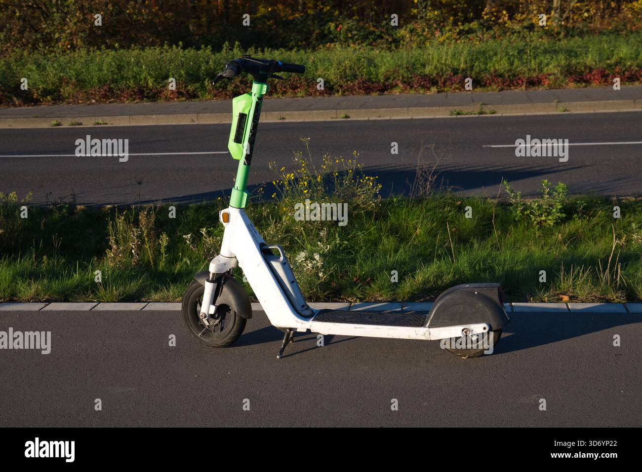 Uno scooter elettrico si trova sul lato di un sentiero asfaltato a Neuss, in Germania. L'impugnatura verde fornisce un contrasto luminoso rispetto al bianco dello scooter f Foto Stock