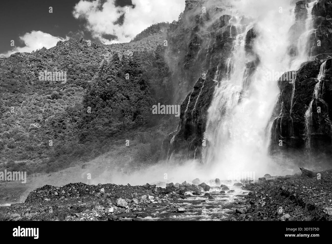 Cascate di Nuranang fiancheggiate da pendii boscosi e creste dell'Himalaya con acqua nebbiosa che precipita verso un fiume sotto il cielo nuvoloso blu nel distretto di Tawang. Foto Stock