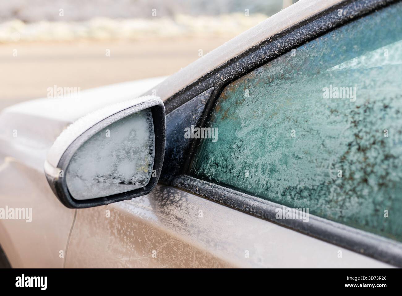 Il ghiaccio ricopre lo specchio laterale e il finestrino di un'auto parcheggiata in un freddo paesaggio invernale. La luce del mattino presto mette in risalto i dettagli ghiacciati e le fredde atmos Foto Stock