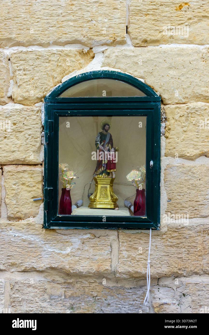 Piccolo Santuario religioso esposto dietro il vetro nella finestra ad arco incastonata nella calda parete di pietra Foto Stock