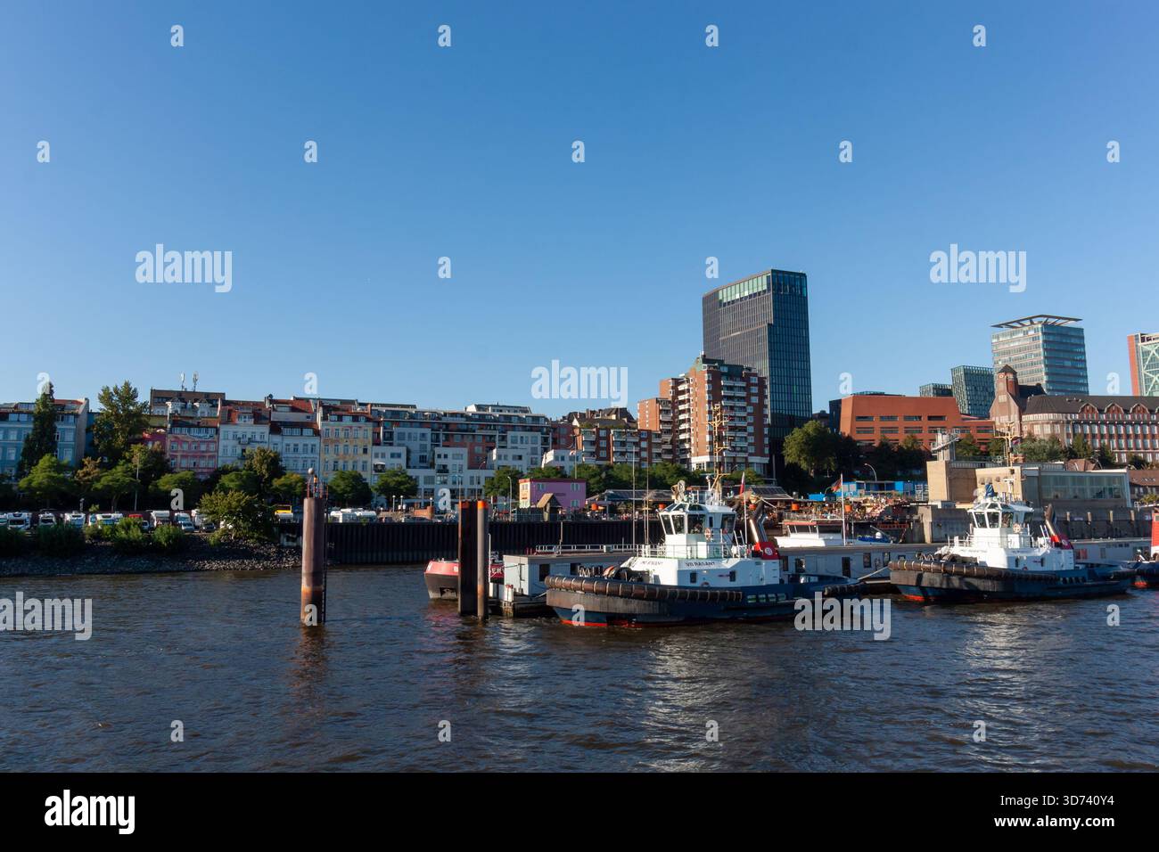 AMBURGO, GERMANIA - 12 AGOSTO 2024: Rimorchiatori attraccati nel porto di Amburgo con lo skyline di St. Pauli e l'Empire Riverside Hotel sullo sfondo sotto un blu Foto Stock