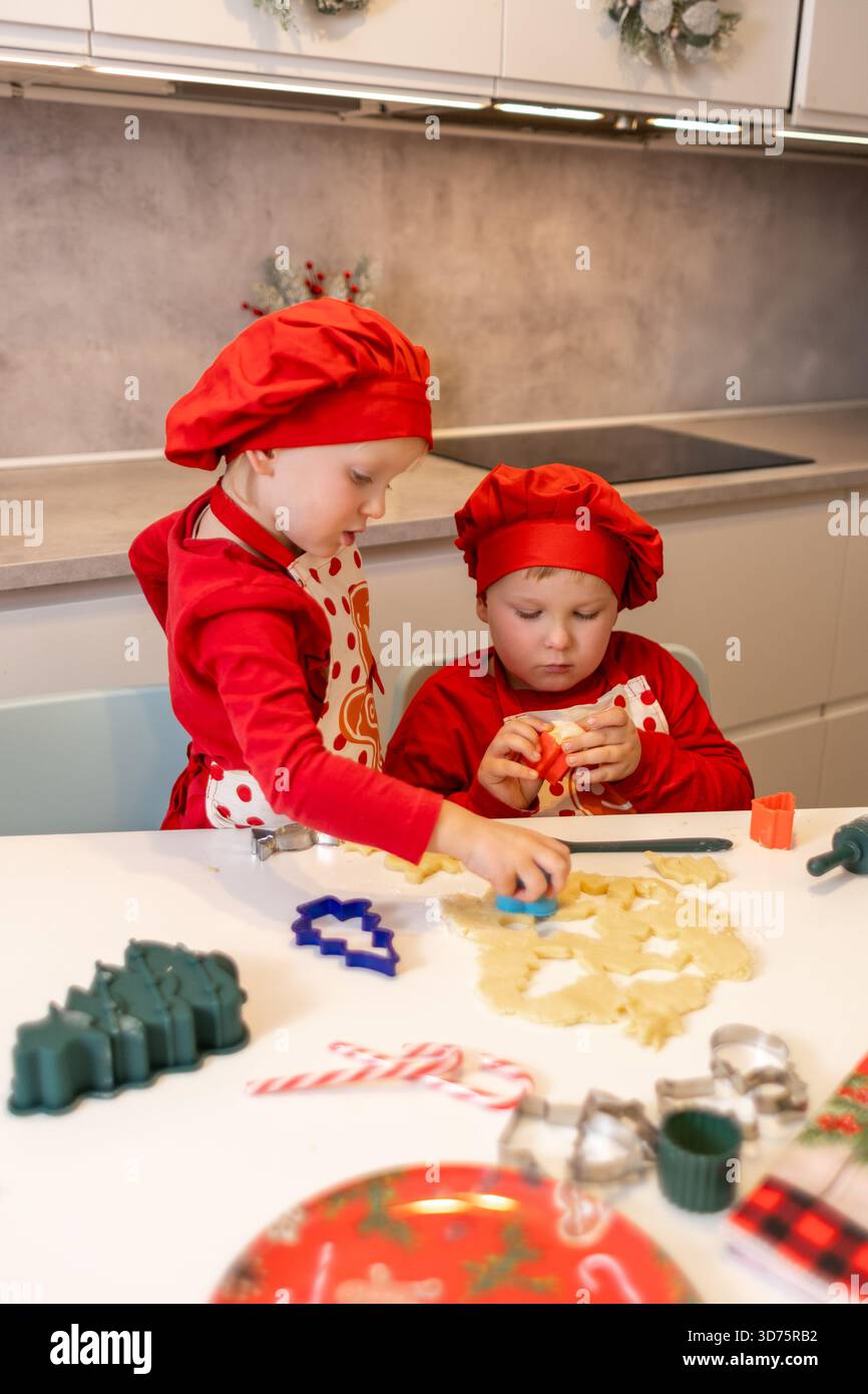 Due bambini preparano con gioia i biscotti di Natale in una cucina calda e decorata. Indossano cappelli da chef rossi abbinati e sembrano concentrati mentre usano il co Foto Stock