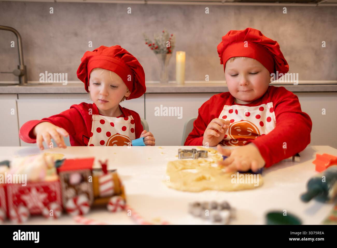 Due bambini allegri in cappelli rossi da chef e grembiuli usano le forchette per preparare l'impasto su un tavolo in una cucina calda. La scena cattura il divertimento di un holida Foto Stock
