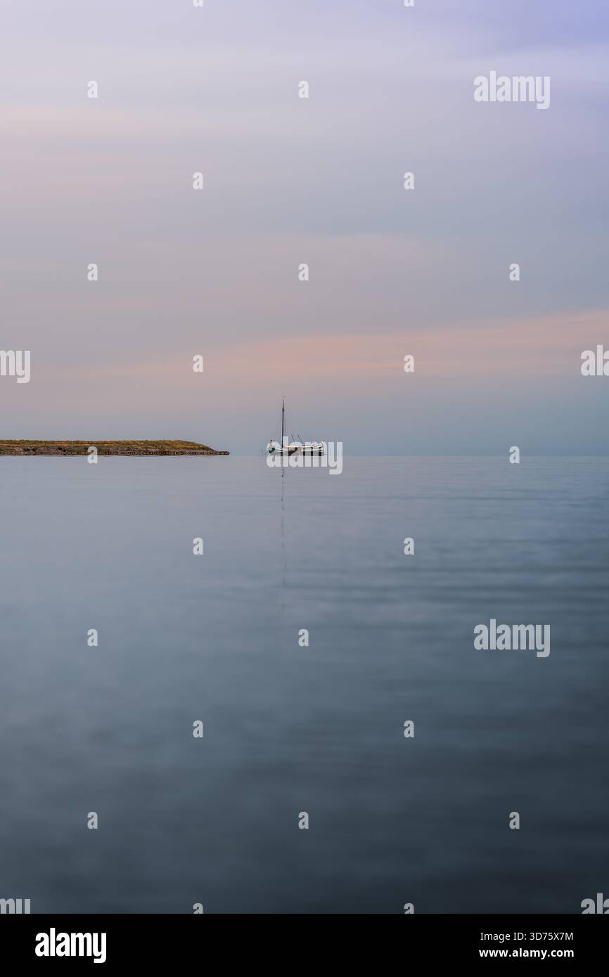 La barca a vela Dutch Tjalk si allontana pacificamente sull'acqua piatta vicino alla riva, con un cielo pastello leggermente nuvoloso al crepuscolo. La scena cattura la serenità serale. Vista ad angolo basso Foto Stock