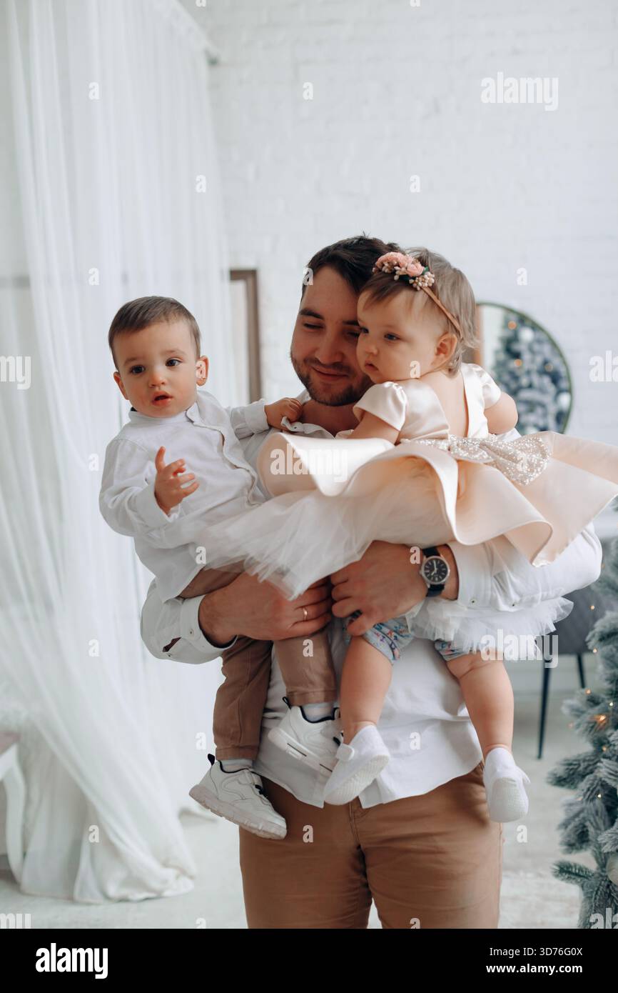 Un padre gioioso porta il figlio e la figlia in un luminoso spazio interno decorato per una festa, creando ricordi caldi di famiglia insieme. Foto Stock