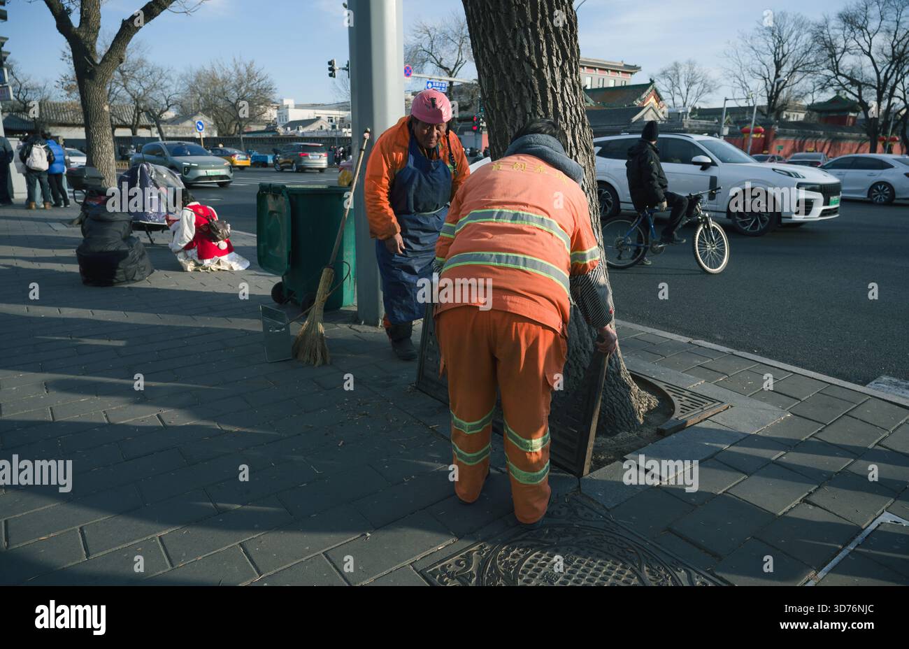 Gli addetti alle pulizie delle strade con uniformi di arancione brillante si aggirano intorno a un albero su un marciapiede invernale affollato a Pechino Foto Stock