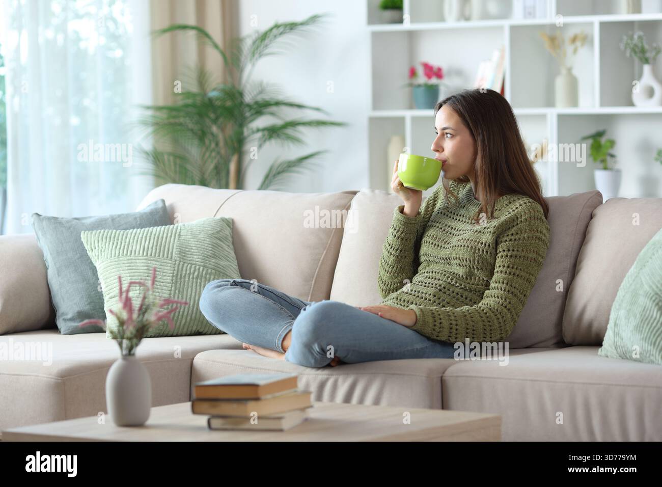 Donna in verde che beve un caffè che guarda lontano seduto su un divano di casa Foto Stock