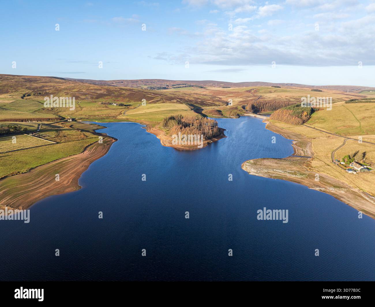 Vista aerea di un tranquillo bacino idrico che rispecchia il cielo, circondato da colline ondulate e campi in sfumature autunnali, una piccola isola boscosa si trova a circa Foto Stock