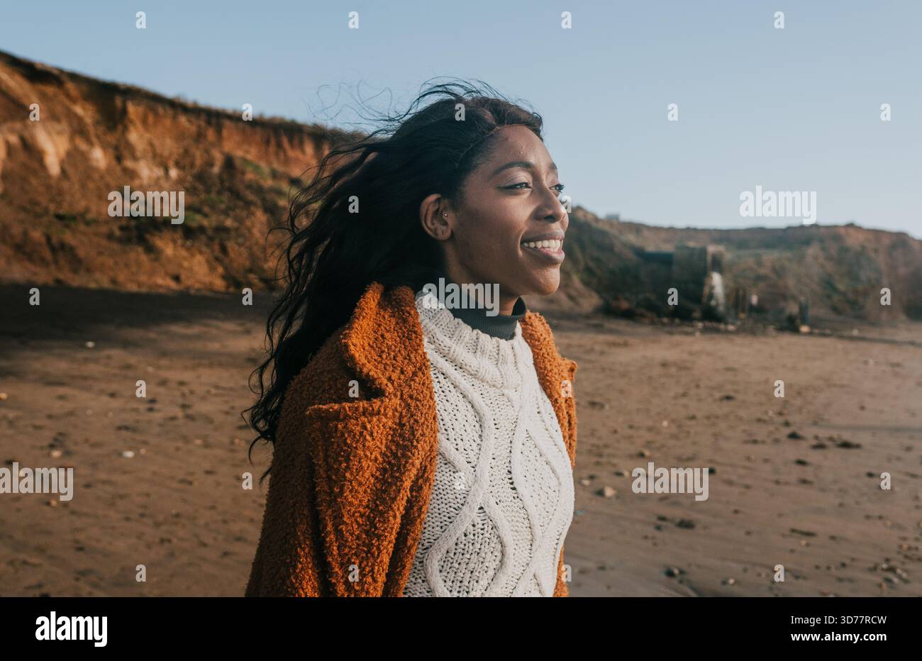 Una donna con i capelli lunghi sorride e indossa un maglione. Lei è in piedi su una spiaggia. La spiaggia è sabbiosa e presenta un'area rocciosa. La donna indossa una W. Foto Stock