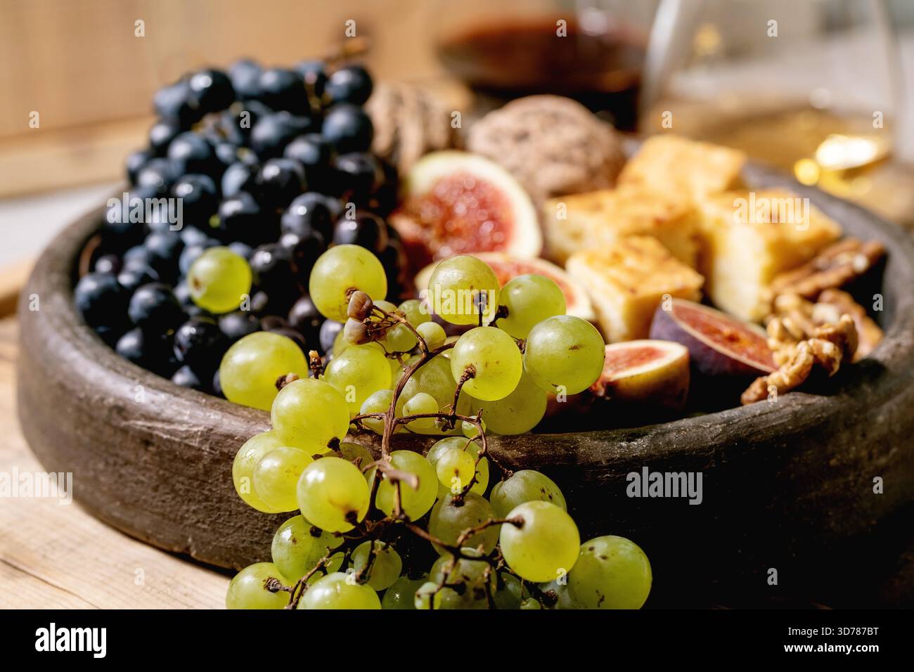 Antipasti con diverse uve, fichi, noci, pane, miele e formaggio di capra su un piatto di ceramica, serviti con bicchieri di vino rosso e bianco su un vecchio sfondo di legno. Primo piano Foto Stock