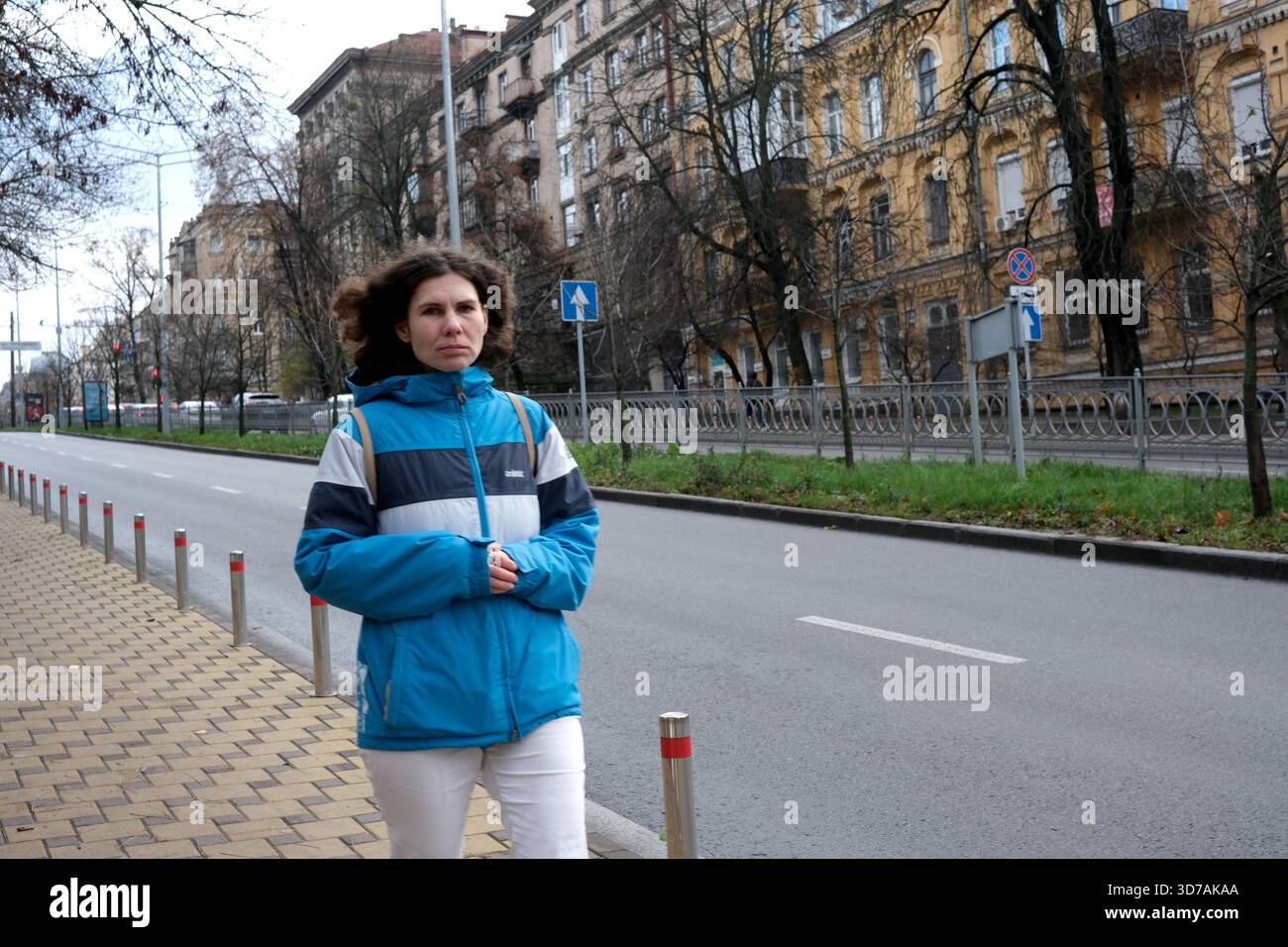 Una donna cammina lungo una tranquilla strada cittadina indossando una giacca calda mentre gli alberi oscillano dolcemente. 24 novembre 2025 Kiev, Ucraina Foto Stock