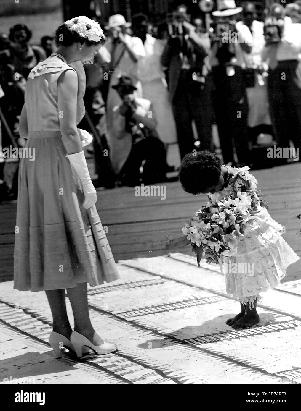 Saluti -- anche il solito autocontrollo della Regina viene scosso mentre la piccola Fijiana, Mei Kainona, 4 anni, la incontra con un bouquet mentre scende a riva alle Fiji, la sua prima tappa nel Pacifico del tour reale. "Che bella", dice mentre Mei si inchina, la presenta con i fiori, le ginocchia sul tappeto di tapa e le batte le mani in un'accoglienza figiana. 3 febbraio 1954. Foto Stock