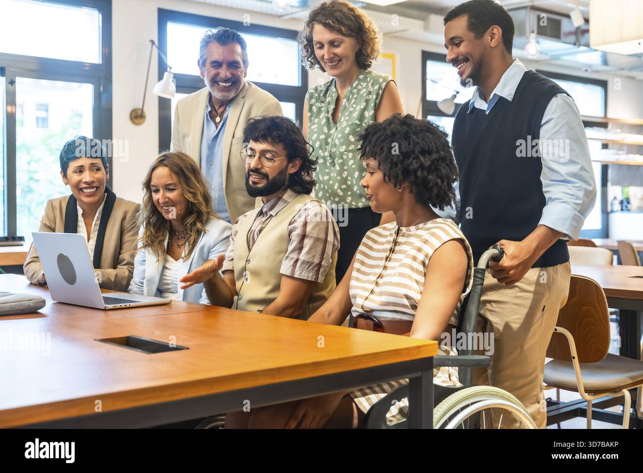 Team aziendale diversificato che sorride e collabora intorno a un notebook in un ufficio di coworking inclusivo, con colleghi con background diversi e un Foto Stock