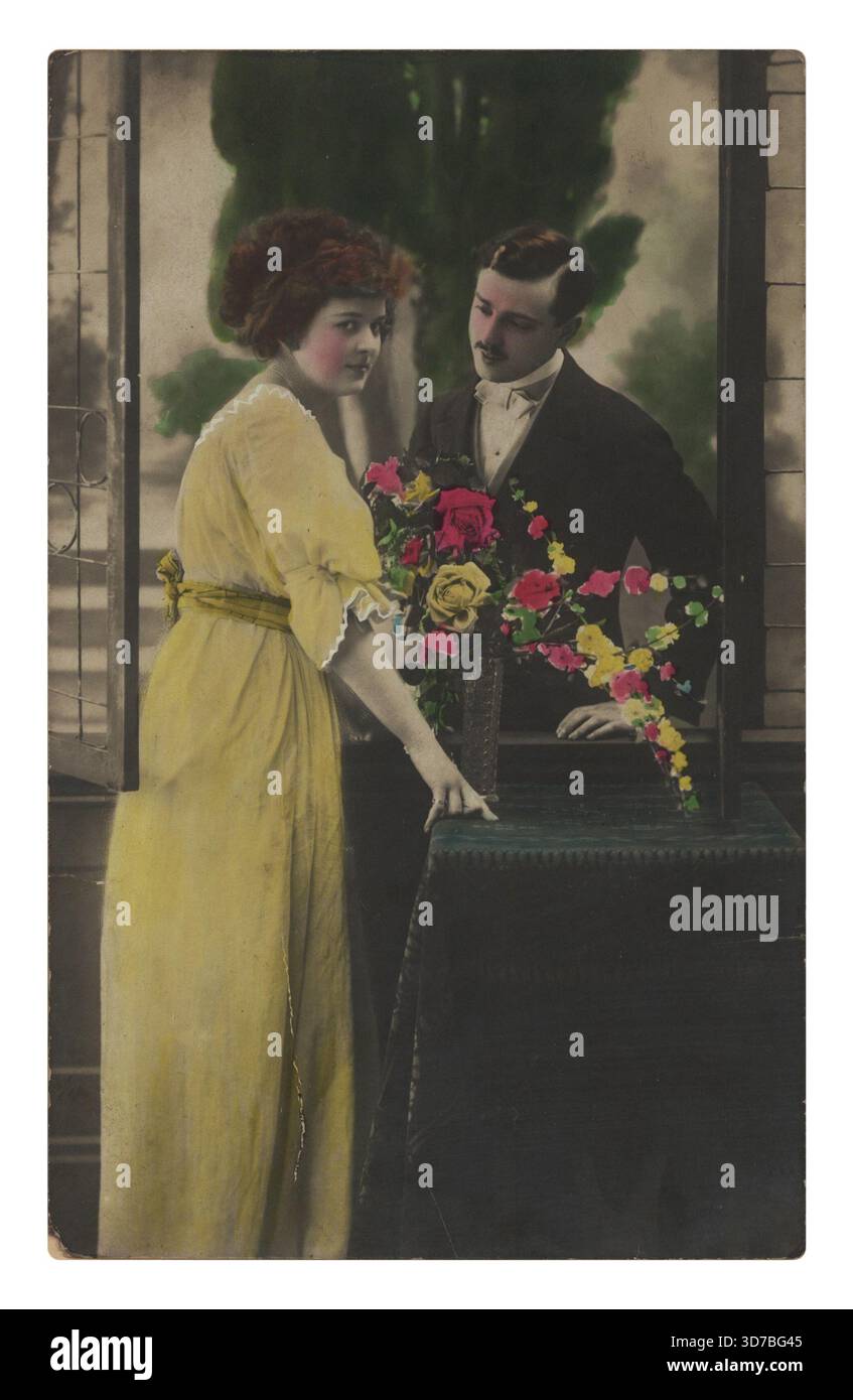 Una donna con un vestito giallo sta accanto a una finestra aperta accanto a un tavolo carico di fiori, mentre un uomo con un vestito la guarda da dietro. Foto Stock